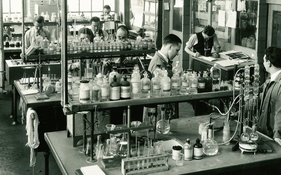 Fotografia antiga do departamento farmacêutico da Sandoz. Foto em preto e branco de uma sala estilo laboratório. Algumas pessoas vestida de jaleco e roupa social estão trabalhando com fracos de vidro dispostos numa mesa de dois níveis, no nível superior existem vários fracos de vidro e na mesa de baixo equipamentos e espaço livre. 