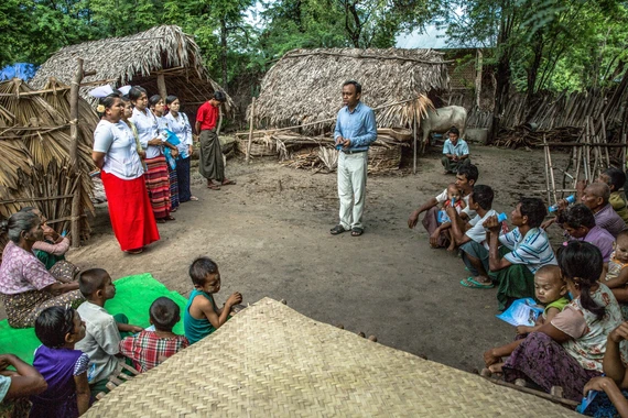 Foto de um local externo rural com um grupo de crianças e adultos atentos a um homem no centro. O ambiente possui estruturas de palha e madeira ao fundo, o chão é de terra, em desfoque atrás estão árvores tropicais. O homem ao centro é negro e veste roupa social azul e bege. As pessoas ao redor são diversas e vestem roupas coloridas como regatas, camisas, shorts e saias. 