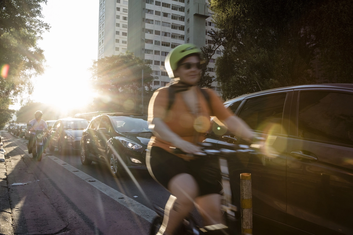 Foto ensolrada de um ambiente externo, com uma moça pilotando bicicleta ao lado de uma rua de carros, prédios e árvores ao fundo. 