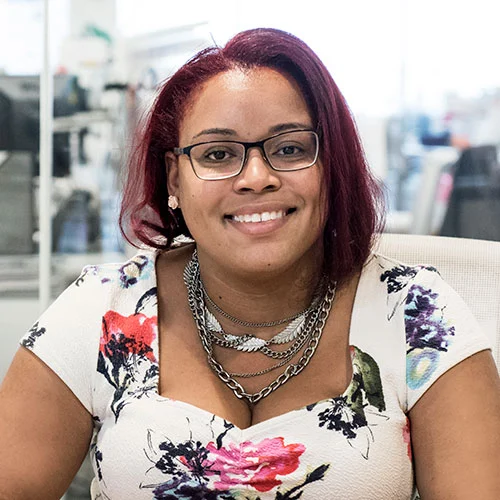 Foto de retrato de uma mulher, morena, do cabelo liso curto vermelho, usa óculos, cordões prata e uma blusa estampada, sorrindo para a câmera. 